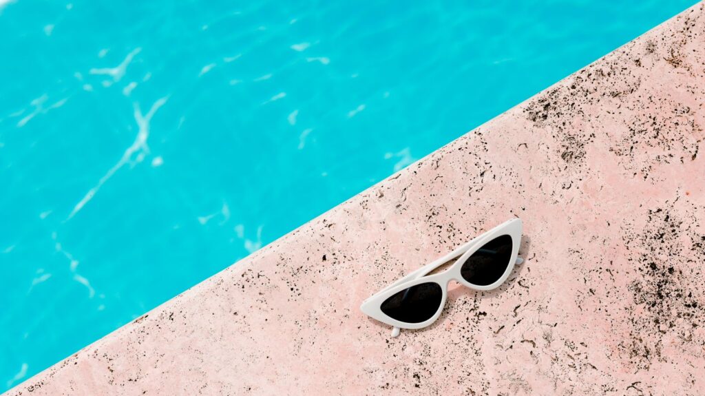 Chic white sunglasses resting on a pink poolside next to clear blue water, perfect for summer vibes.